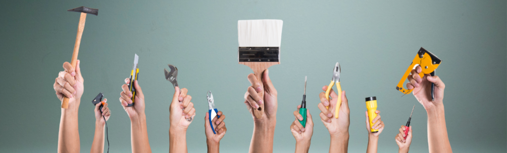 Hands holding various tools against a gray background, featuring a hammer, paintbrush, pliers, and screwdriver.