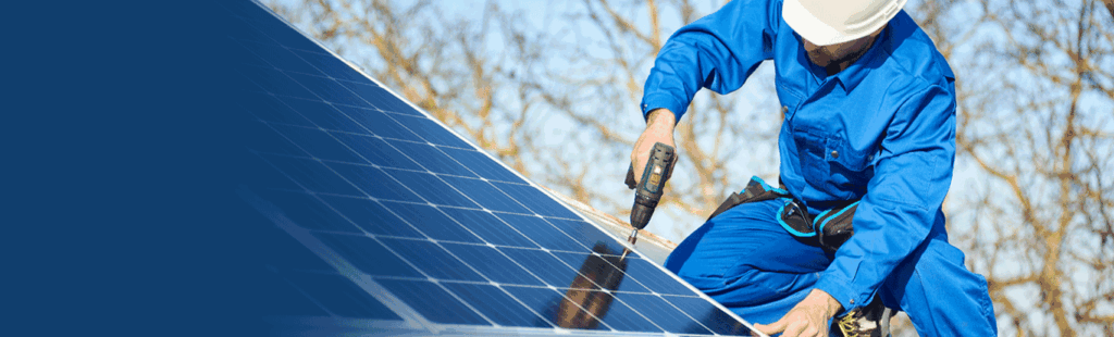 Worker installing solar panels, wearing blue uniform and hard hat, with a drill in hand against a clear sky background.