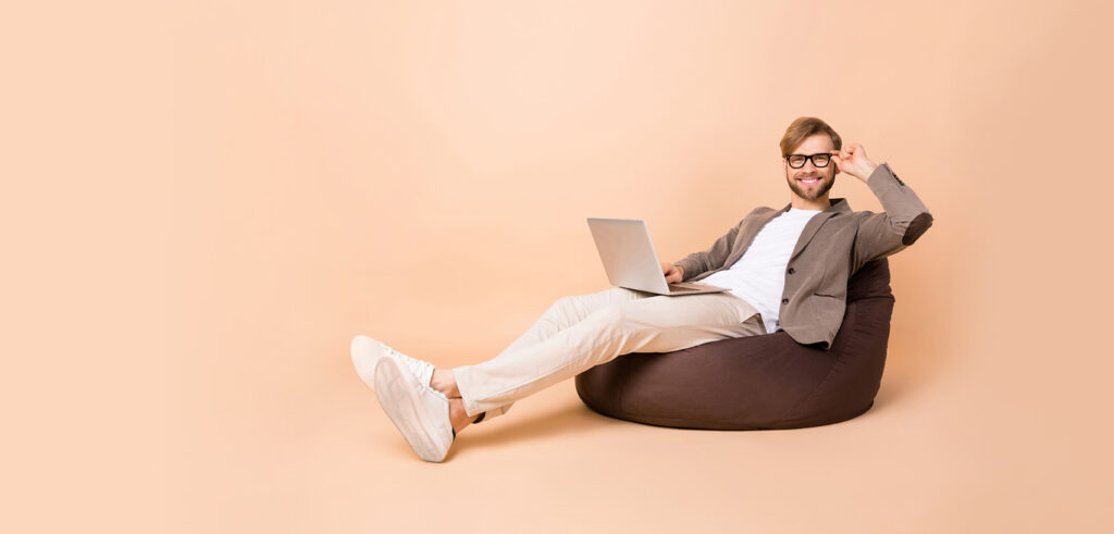 Man sitting on bean bag with laptop, wearing glasses and casual attire, smiling against a neutral background.