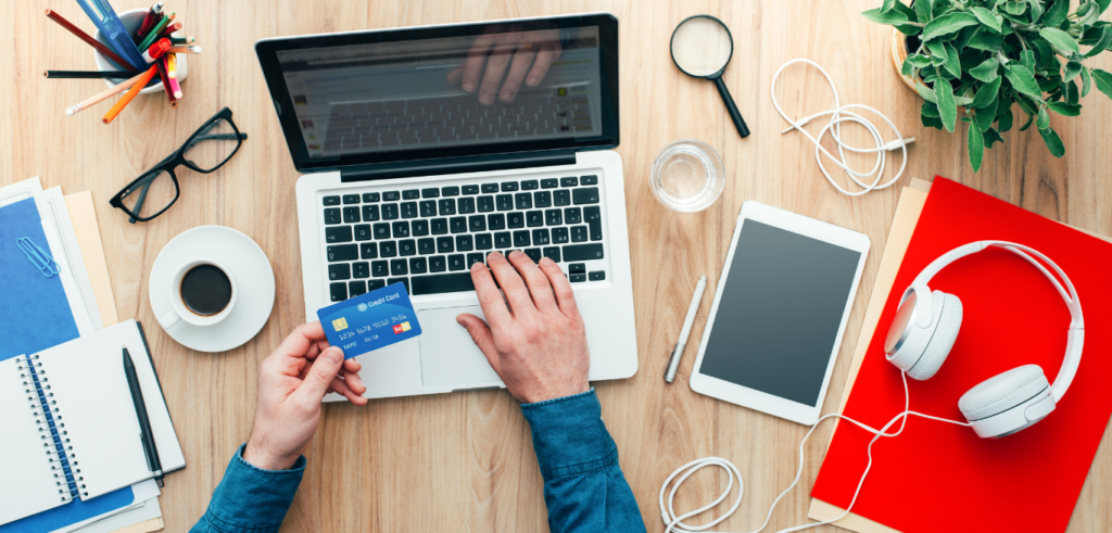 Person shopping online with laptop and credit card, surrounded by office supplies on a wooden desk.