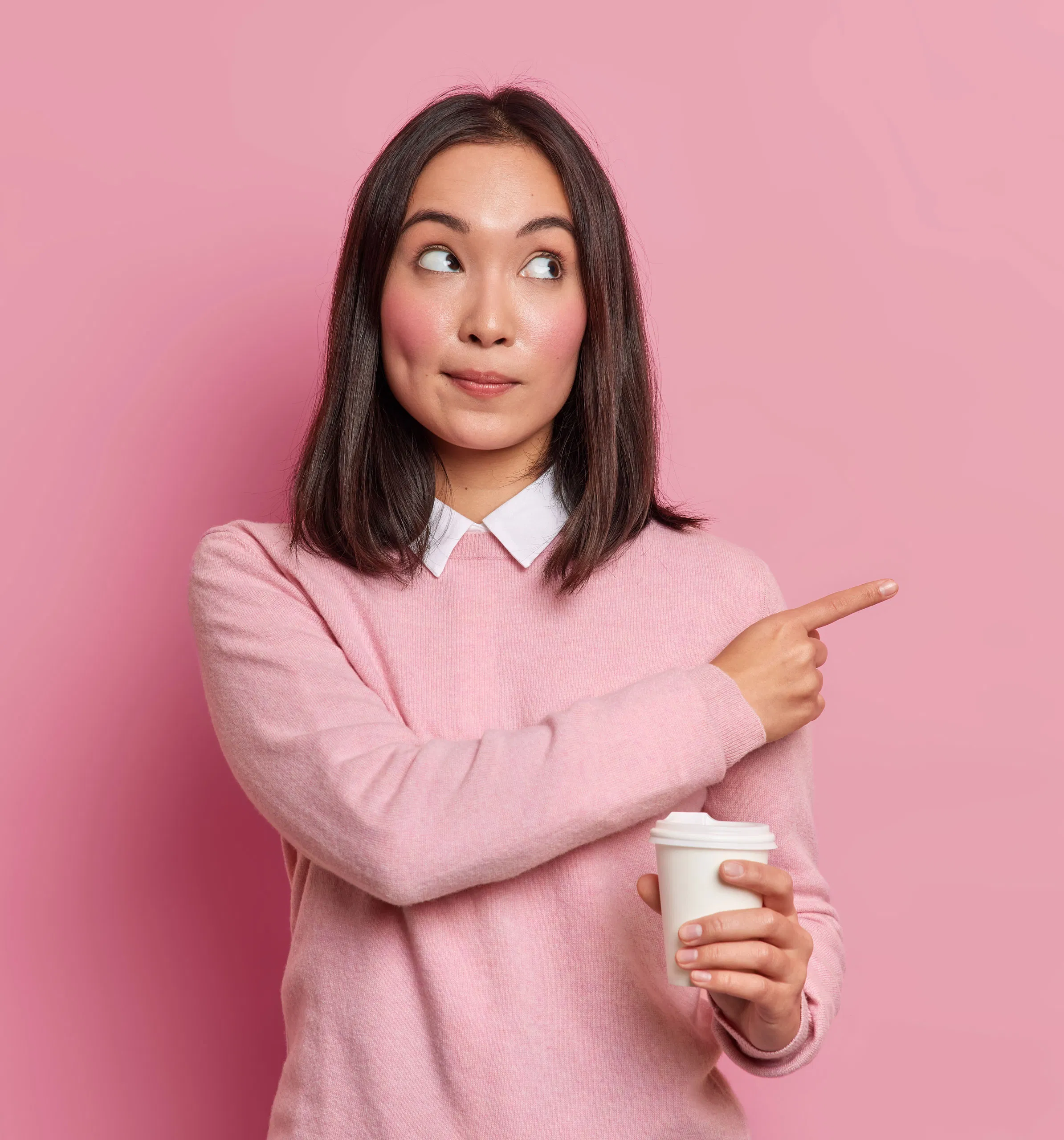 Woman in pink sweater with a coffee cup playfully points sideways against a pink background.
