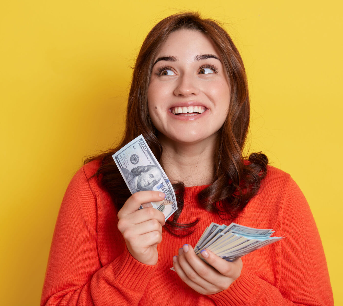 Woman in orange sweater holding cash with a joyful expression on a yellow background.