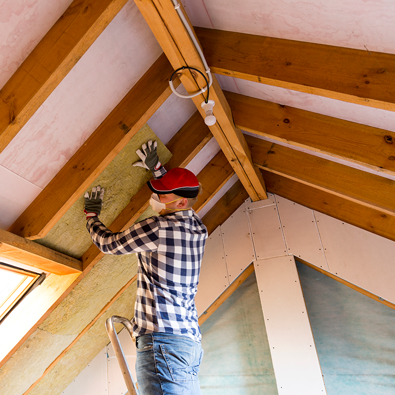 Man installing attic insulation, wearing safety gear, on wooden beams for energy efficiency.