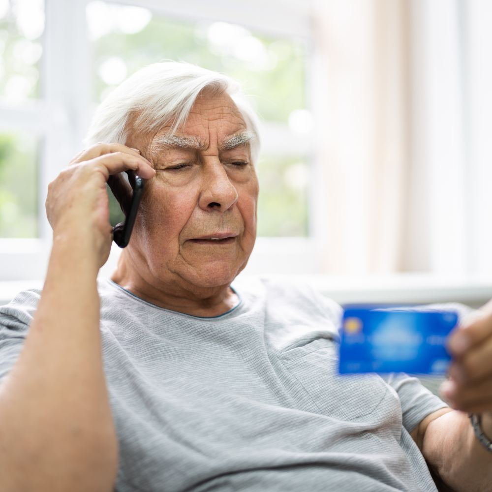 Senior man on phone holding credit card, seated indoors by window.