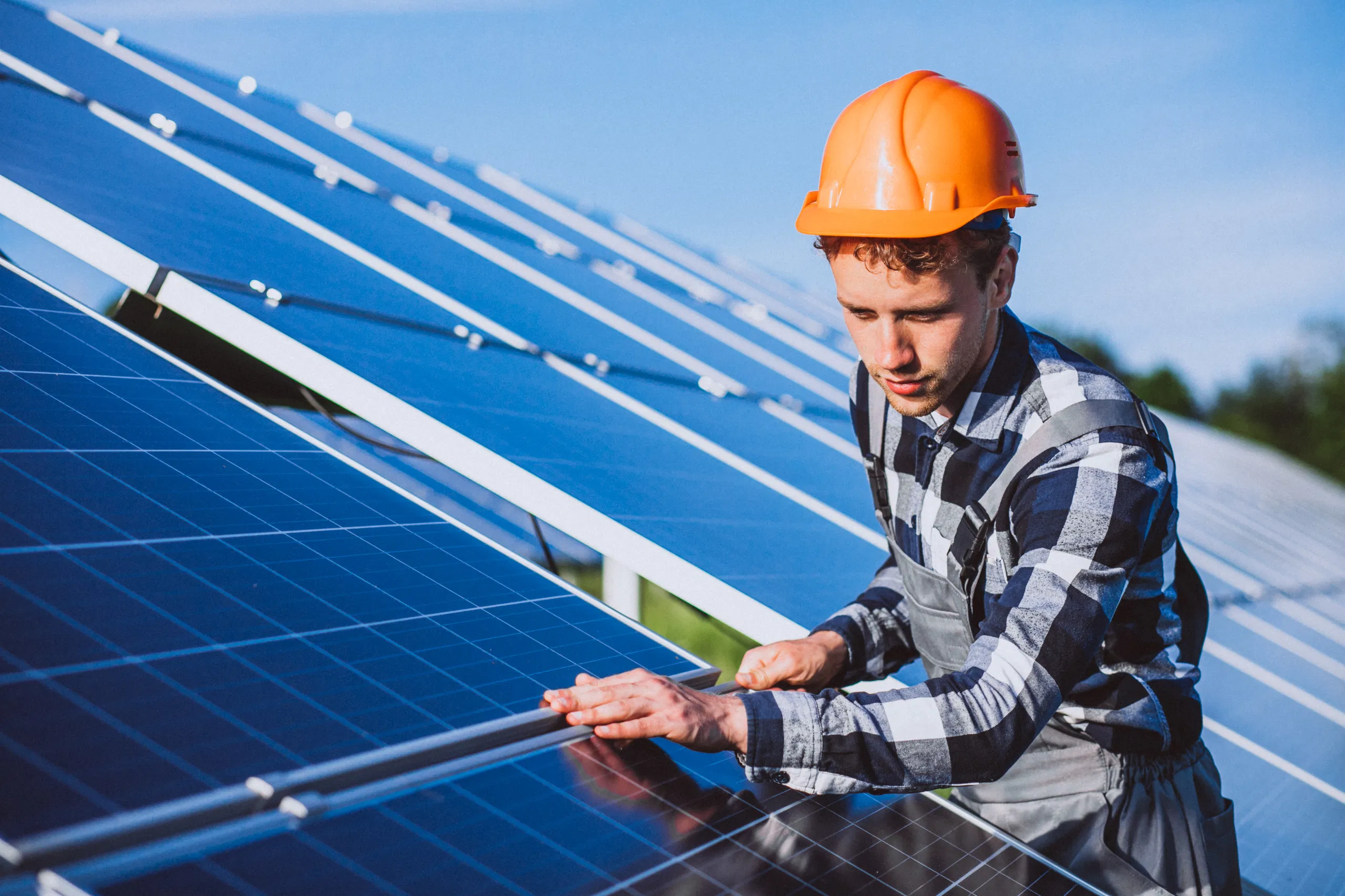 Technician inspecting solar panels in sunny field, wearing safety helmet and uniform.