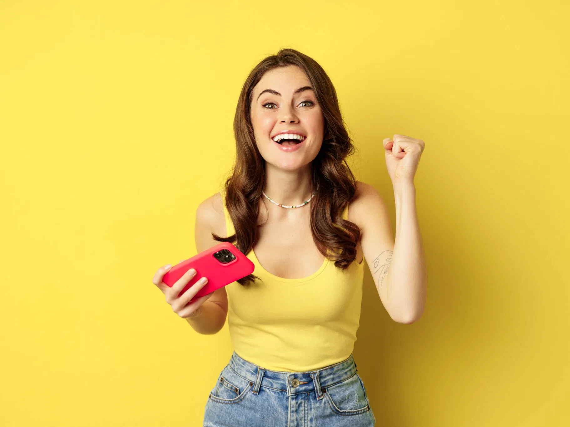 Young woman celebrating with a pink phone, yellow background, wearing a yellow top and jeans.