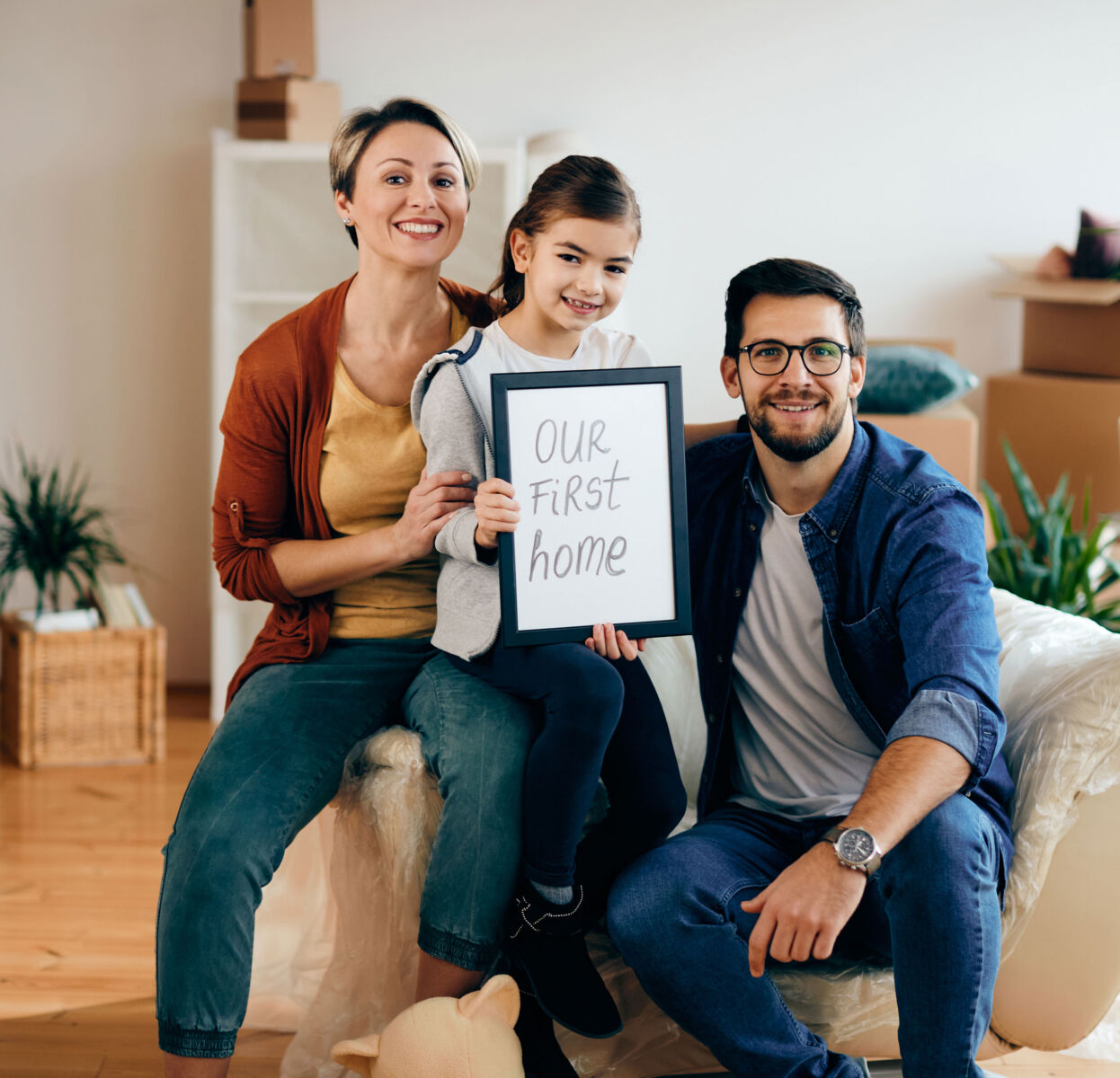 Family celebrating their first home purchase, holding a sign that reads Our First Home surrounded by moving boxes.