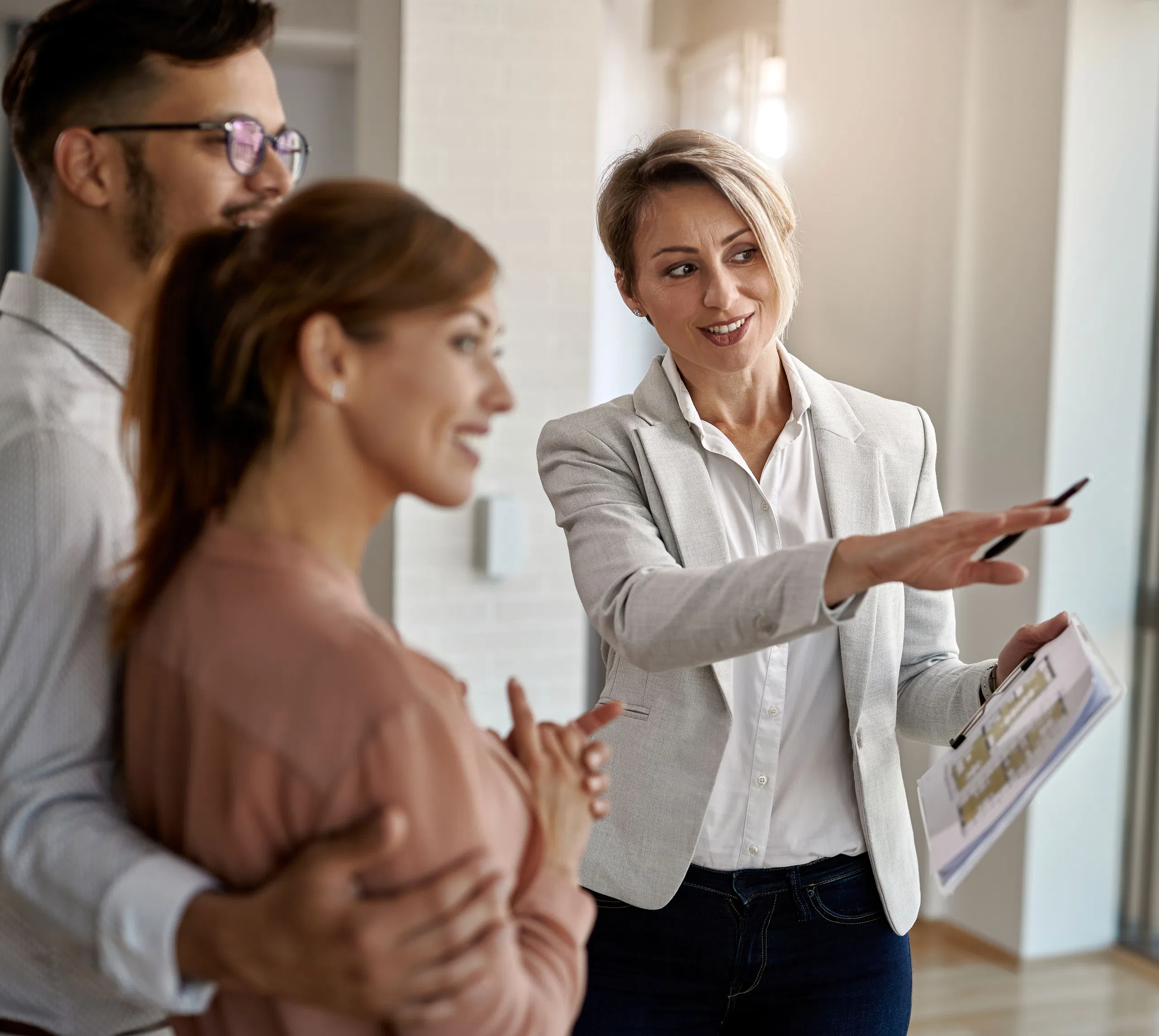 Real estate agent showing happy couple new property, discussing features, and holding a clipboard inside a bright home.
