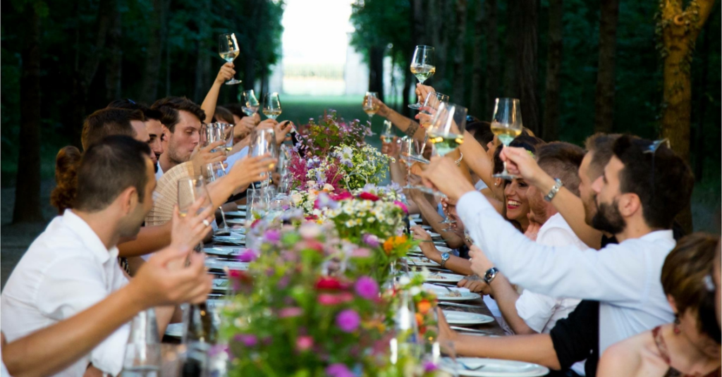 Outdoor group dinner at long table with people raising glasses in toast, surrounded by vibrant flowers.