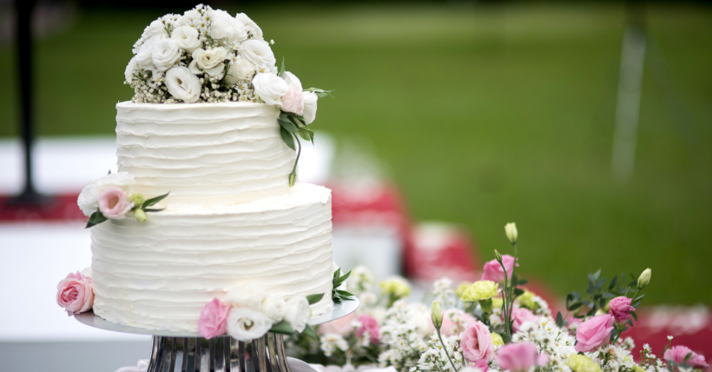 Elegant two-tier white wedding cake with floral decorations on a vibrant outdoor background.