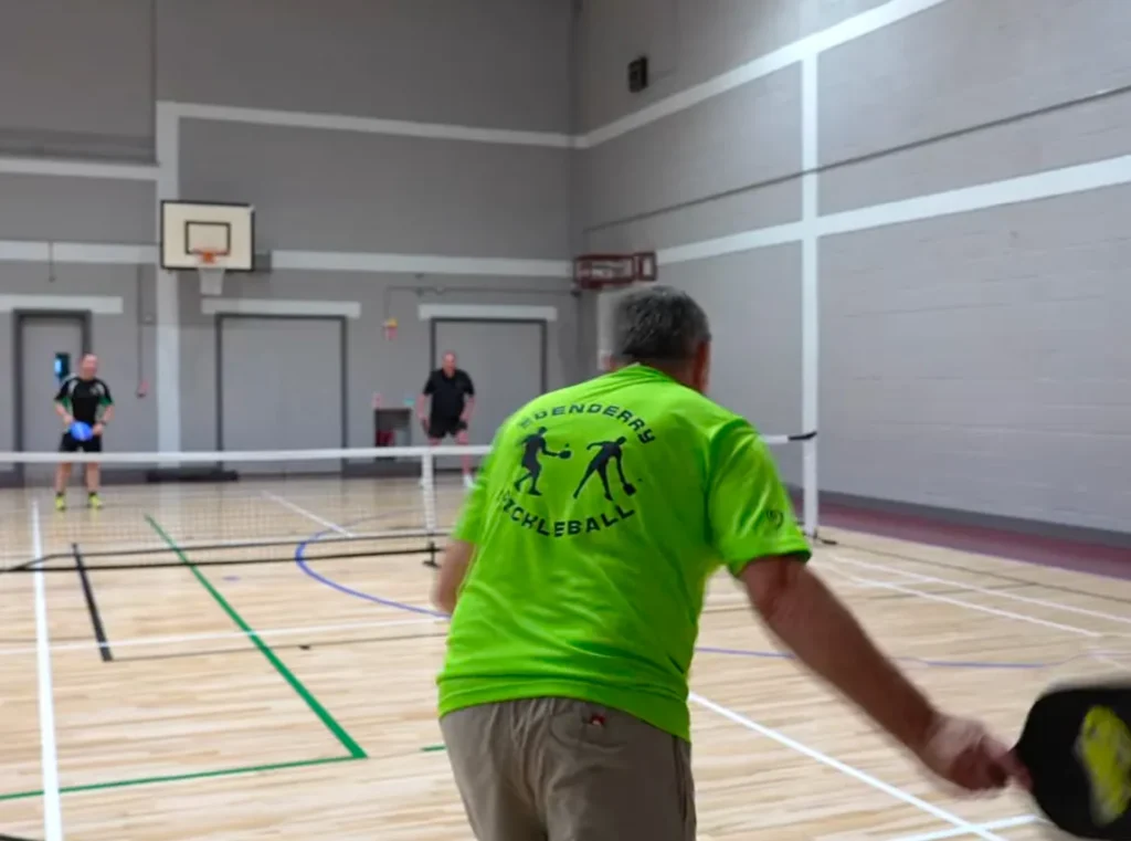 Men playing pickleball indoors, wearing bright green club shirts, focused on the game at a sports facility.