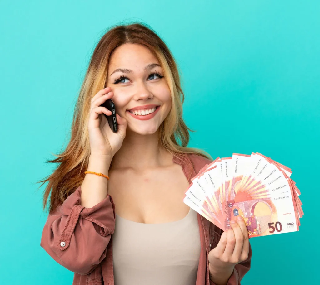 Woman smiling on phone, holding euro bills in hand against turquoise background, depicting financial success and positivity.
