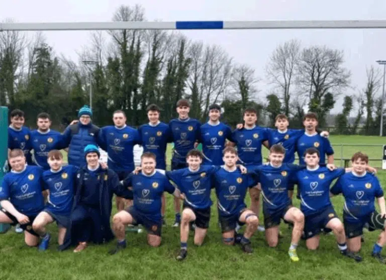 Rugby team in blue uniforms posing on a field with goal posts and trees in the background.