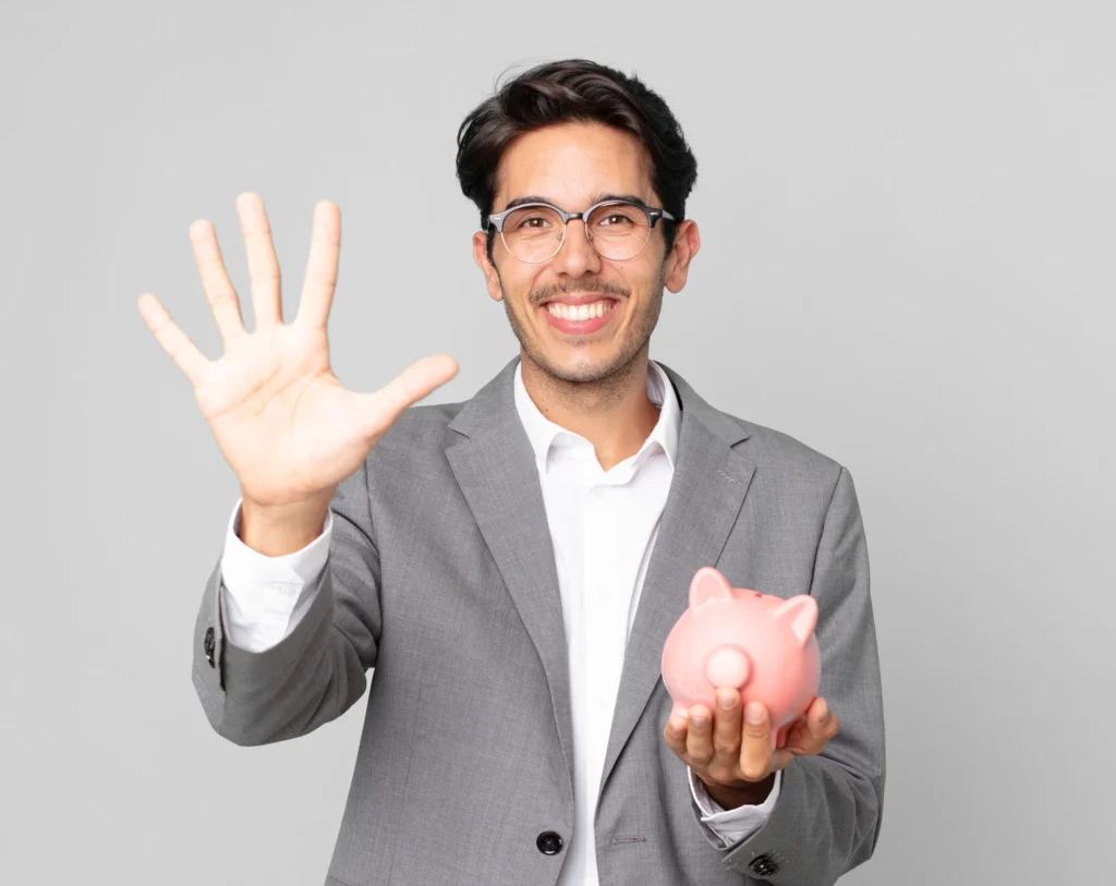 Smiling man in grey suit holding piggy bank, waving with five fingers, on plain background.