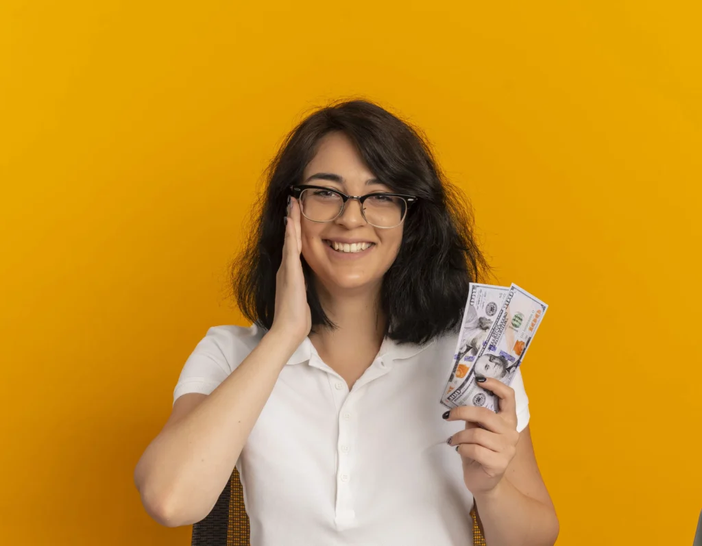 Smiling woman holding dollar bills against a vibrant yellow background, symbolizing wealth and financial success.
