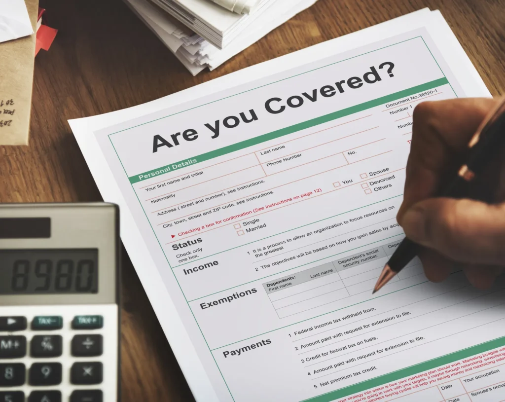 Person completing insurance coverage form on wooden desk with calculator and documents.