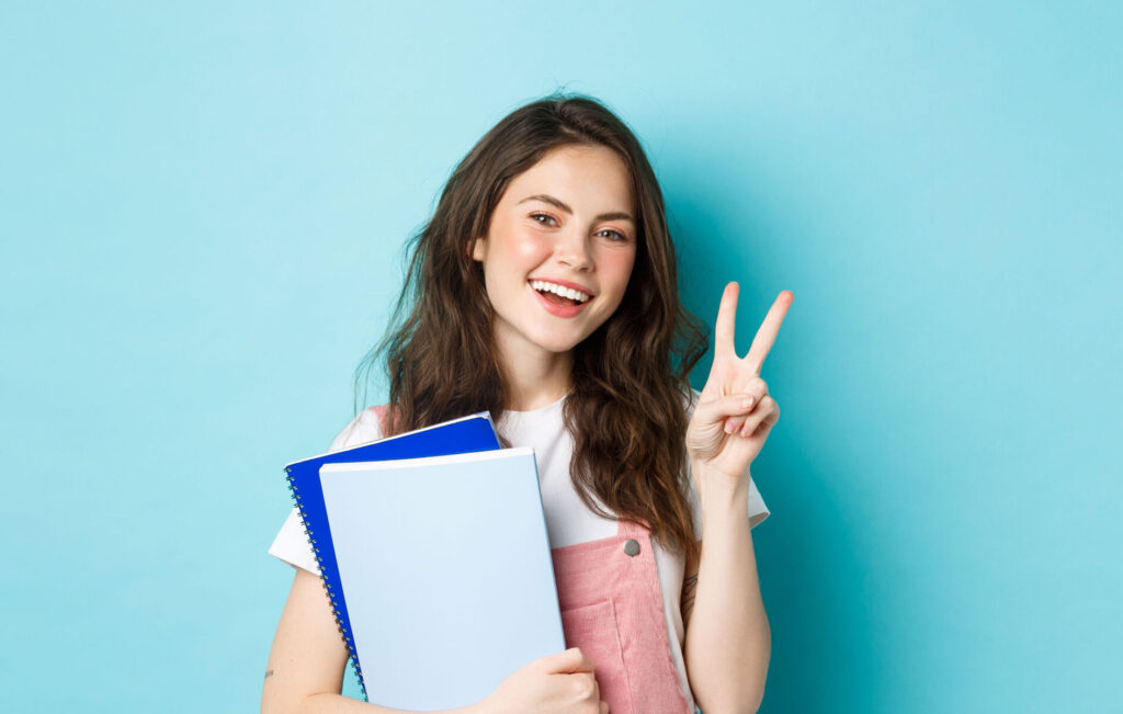 Young woman smiling with notebooks, making a peace sign against a blue background.