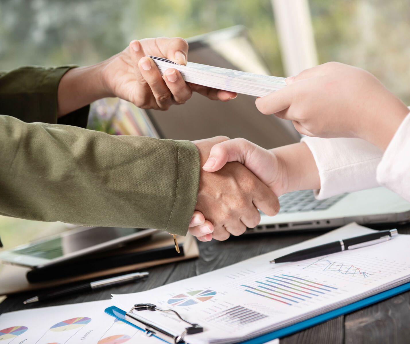 Business handshake with document exchange over desk with charts, conveying partnership and financial collaboration.