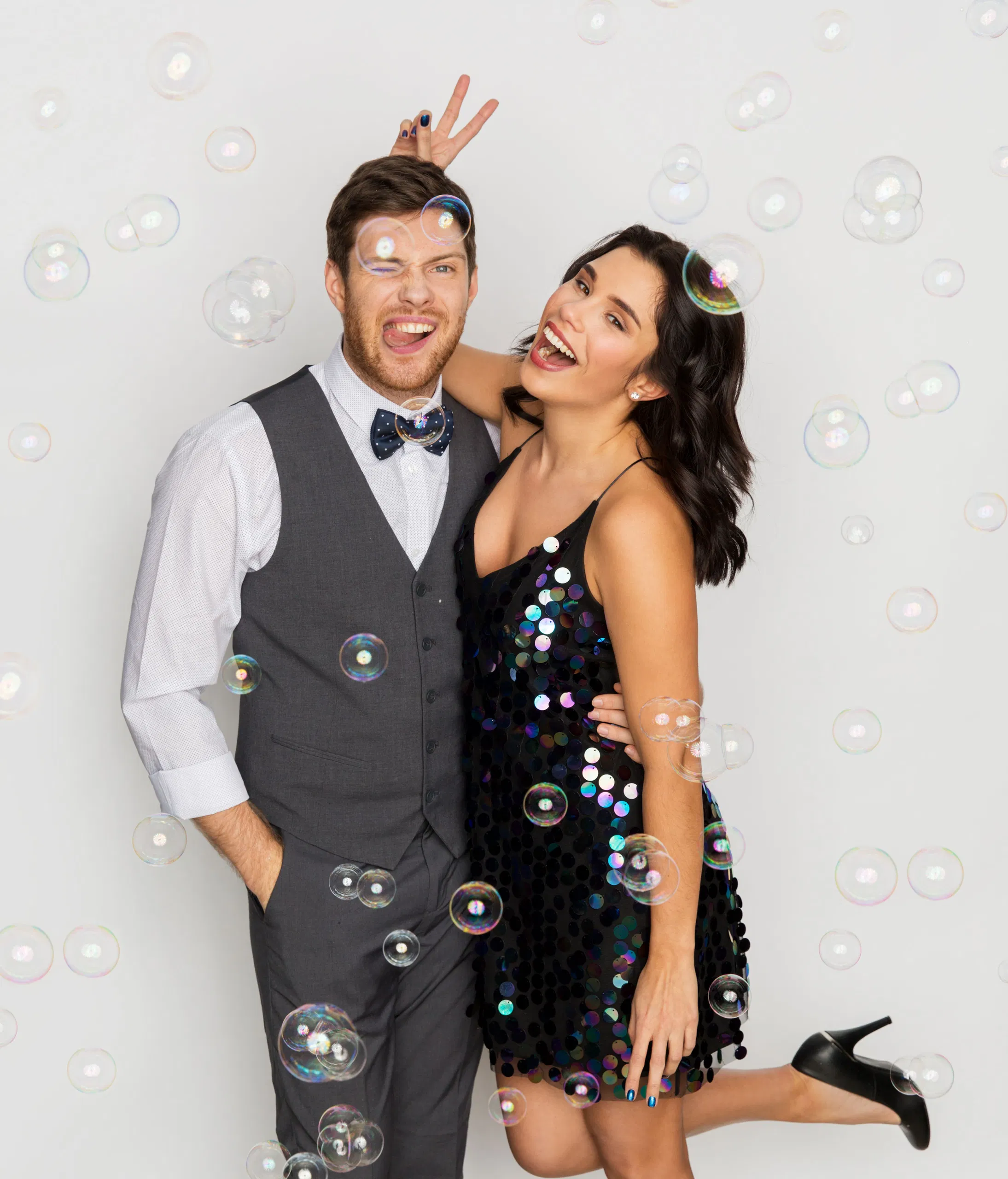 Couple posing playfully with bubbles, man in grey vest winking, woman in black sequin dress smiling.