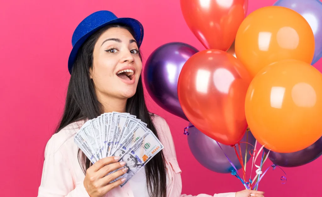 Woman celebrating with balloons and money on a vibrant pink background, wearing a blue hat.