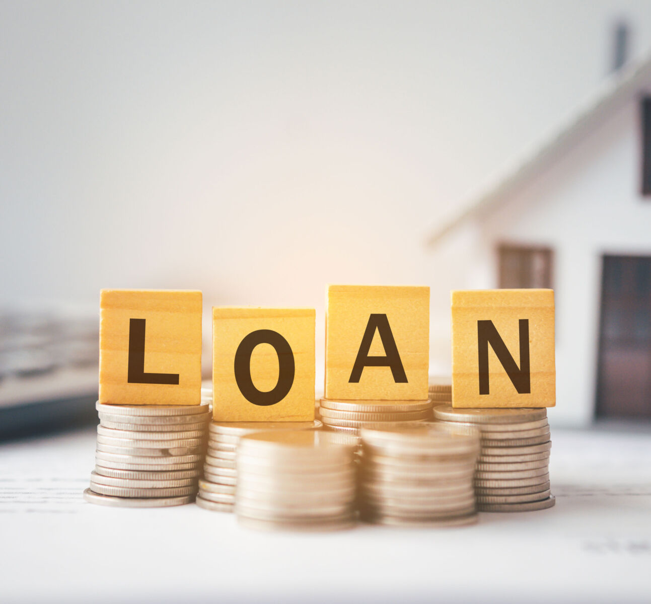 Wooden blocks spelling LOAN on stacked coins, with blurry house background, symbolizing mortgage or home financing.
