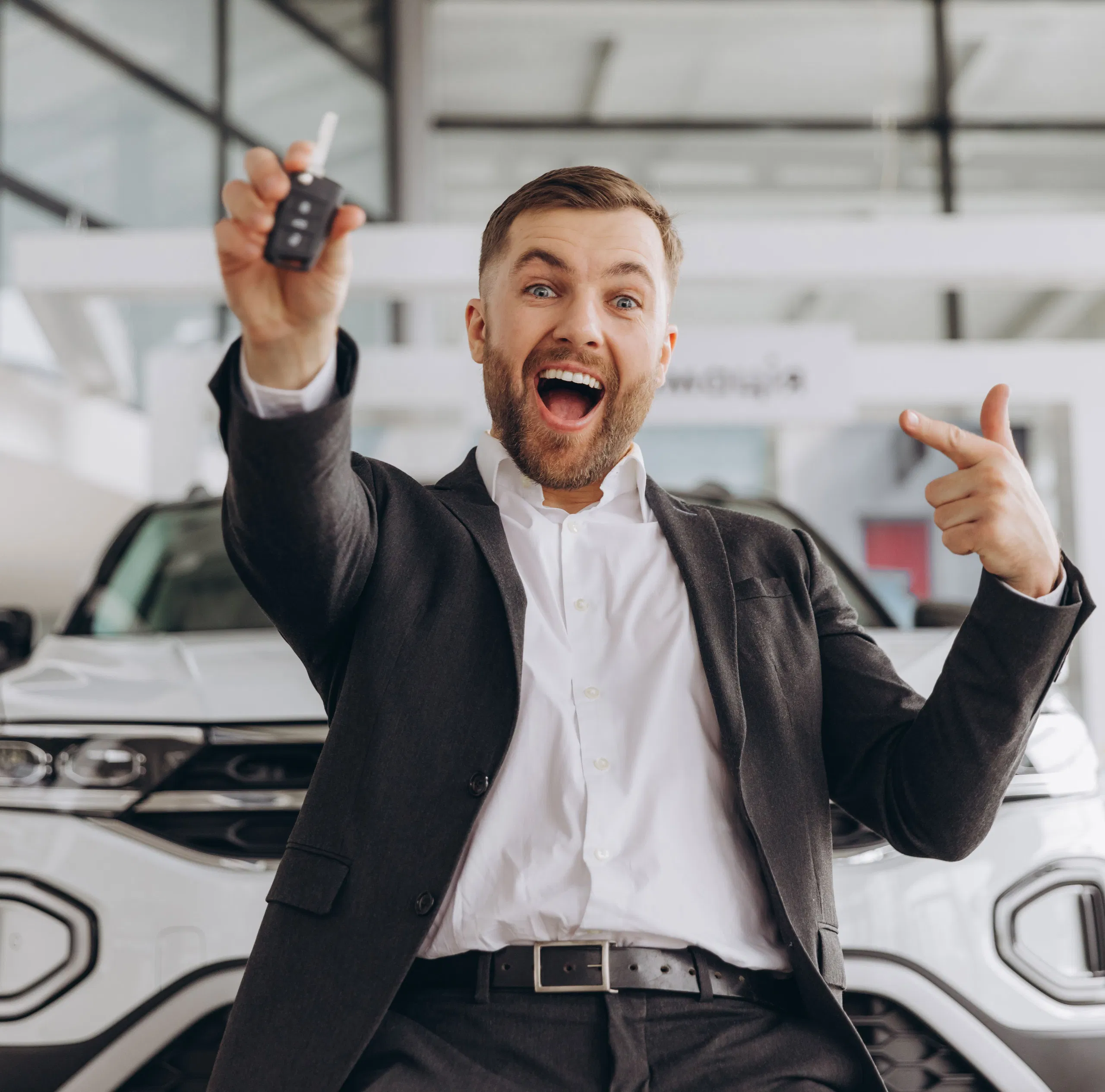 Man celebrating new car purchase, smiling and pointing at car keys in dealership showroom.