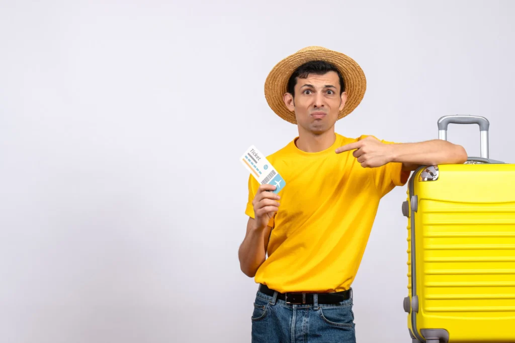 Man in yellow shirt and straw hat holding a plane ticket, pointing at his yellow suitcase with a confused expression.