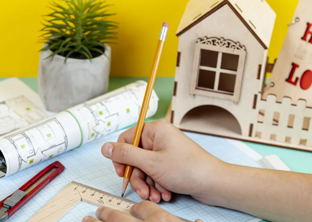 Person drawing architectural plans with ruler and pencil, next to model house and plant.