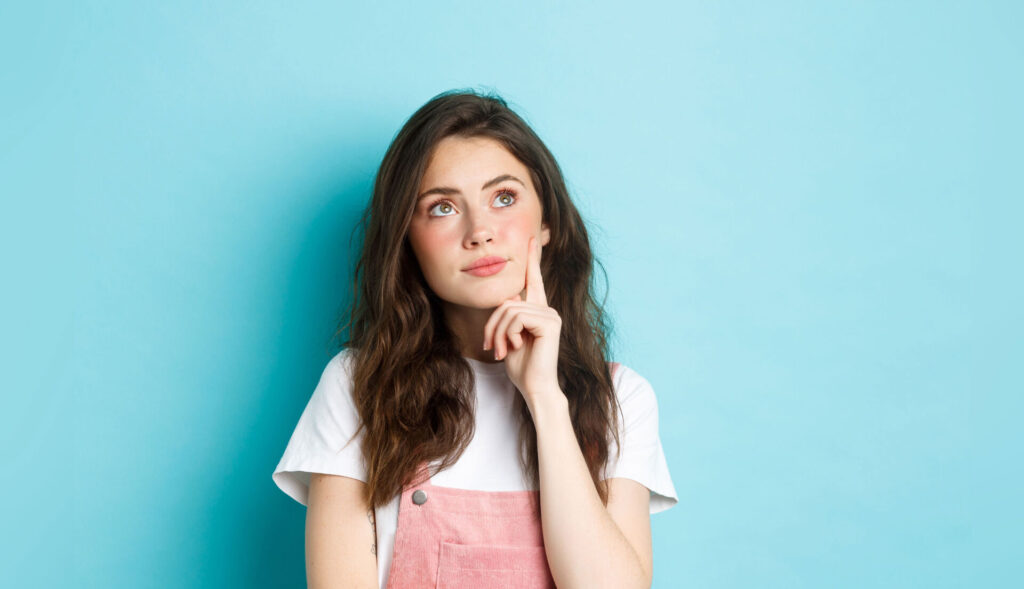 Woman pondering against blue background, wearing a pink jumper and white shirt, thinking concept.