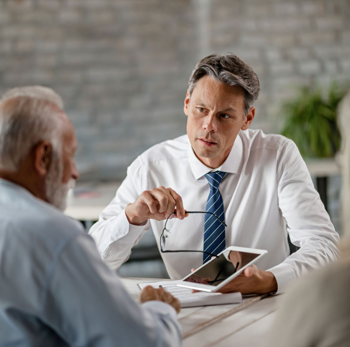 Business meeting between two men discussing over a tablet, one holding glasses, in a modern office setting.