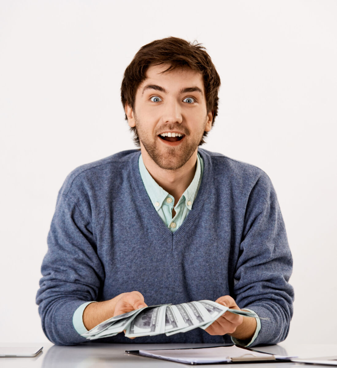 Excited man holding cash fan, wearing blue sweater, sitting at a table with notebook.