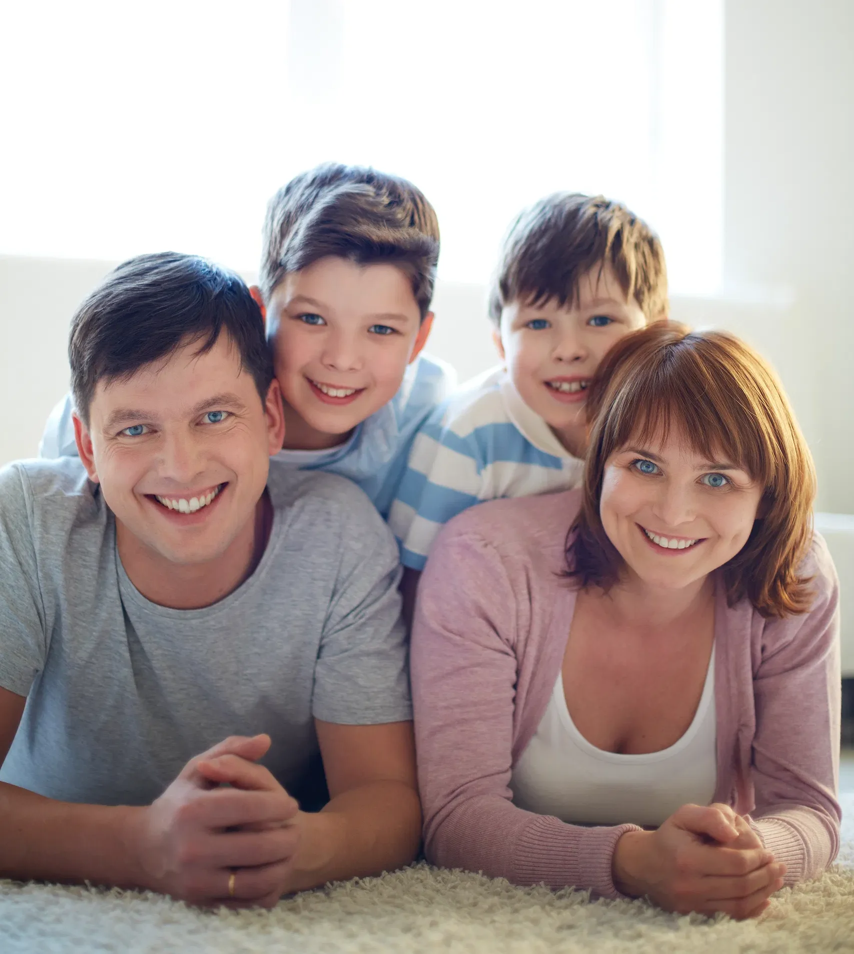 Happy family of four enjoying time together at home, smiling while lying on a soft carpet in a cozy living room.