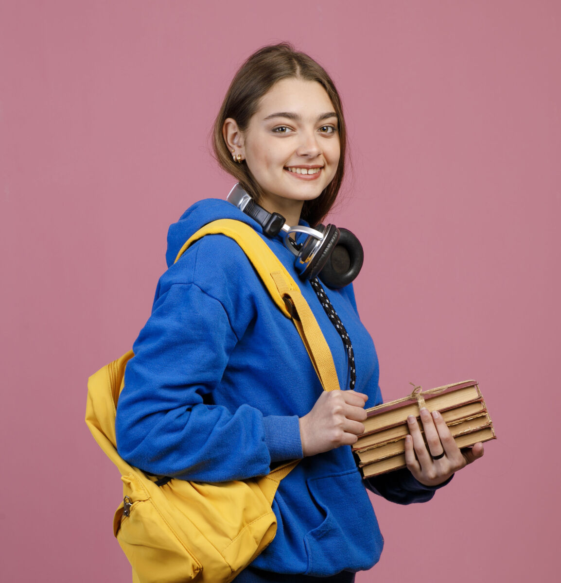Smiling student in a blue hoodie with yellow backpack, headphones, and books against a pink background.