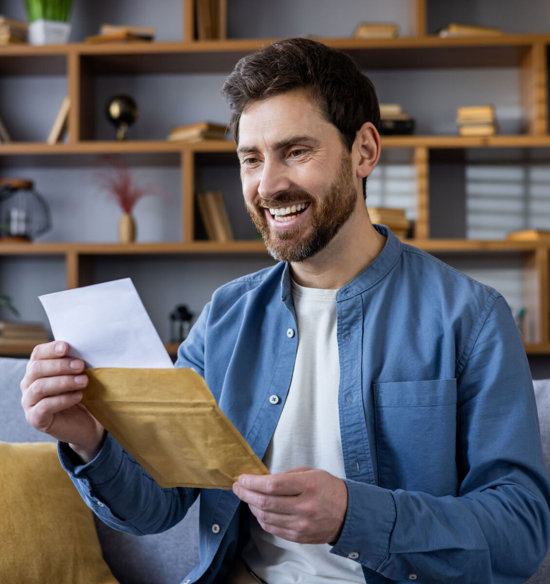 Man smiling at letter received in envelope while relaxing on sofa at home.