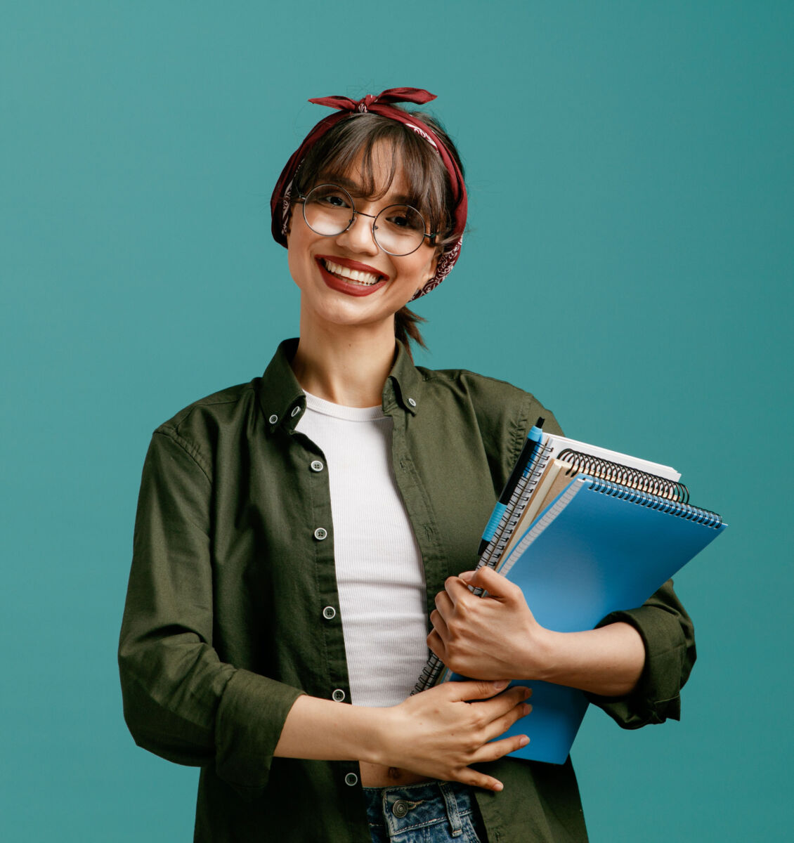 Smiling person with glasses holding notebooks, wearing a red headband and green shirt against a teal background.