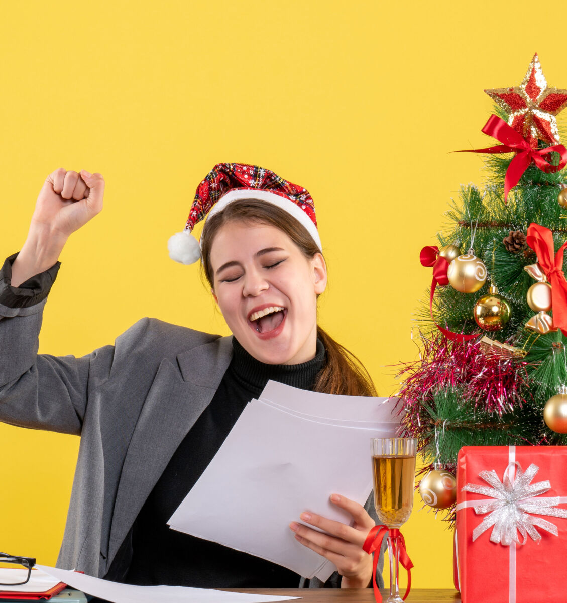 Happy woman in Santa hat celebrating with papers and Christmas tree, wearing a suit, against a yellow background.