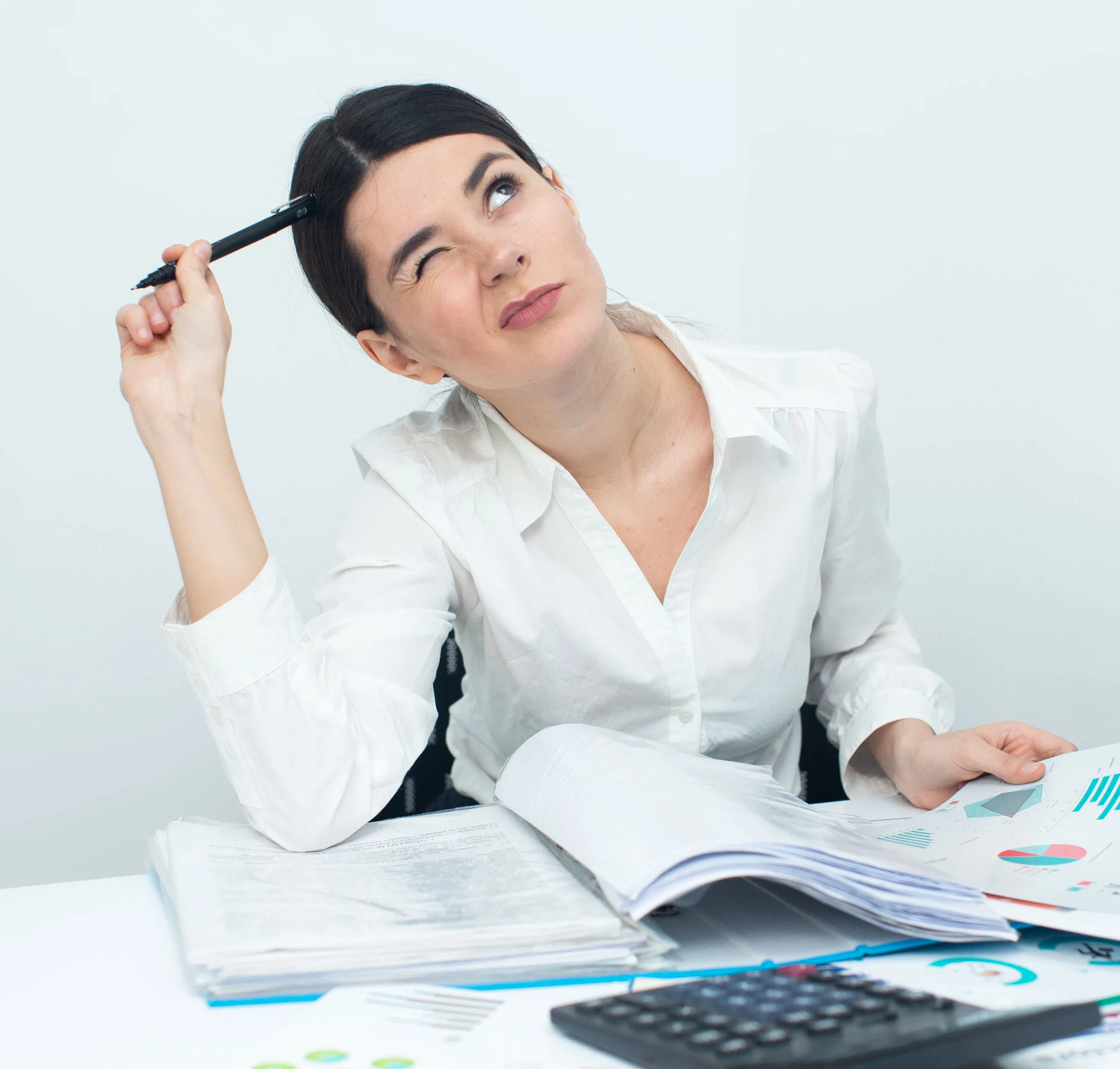 Woman in white shirt pondering over documents, holding pen to head, with calculator on desk.