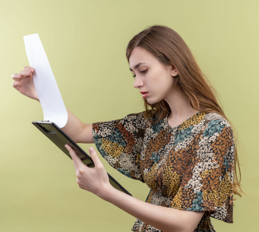 Woman reading a document on a clipboard, focused expression, multicolored dress, neutral background.