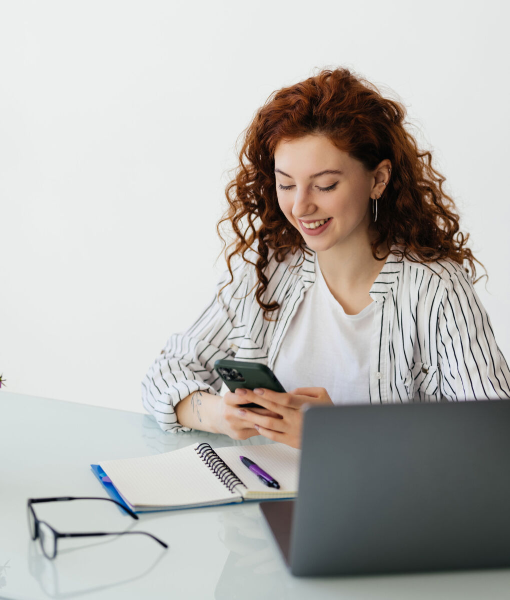 Woman smiling while using smartphone at desk with notebook and laptop.