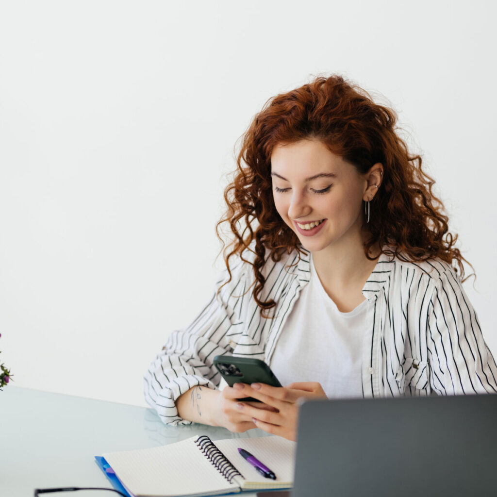 Smiling woman using smartphone at desk with laptop and notebook, wearing striped blouse against white background.